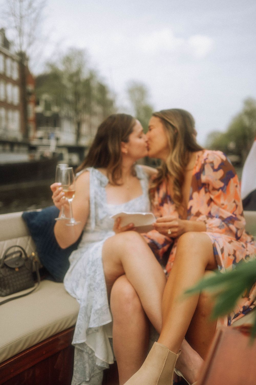 Two women sharing a kiss on a boat cruise, holding champagne glasses, wearing summer dresses.