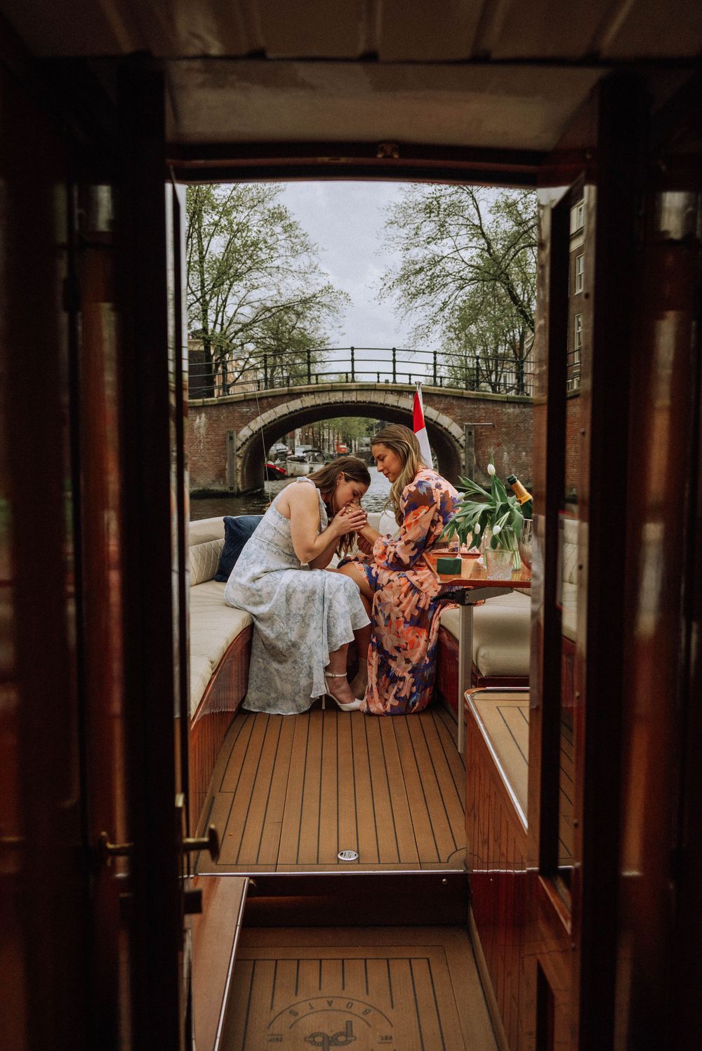 Two women sharing a moment on a classic wooden canal boat in Amsterdam, with a stone bridge visible in the background.