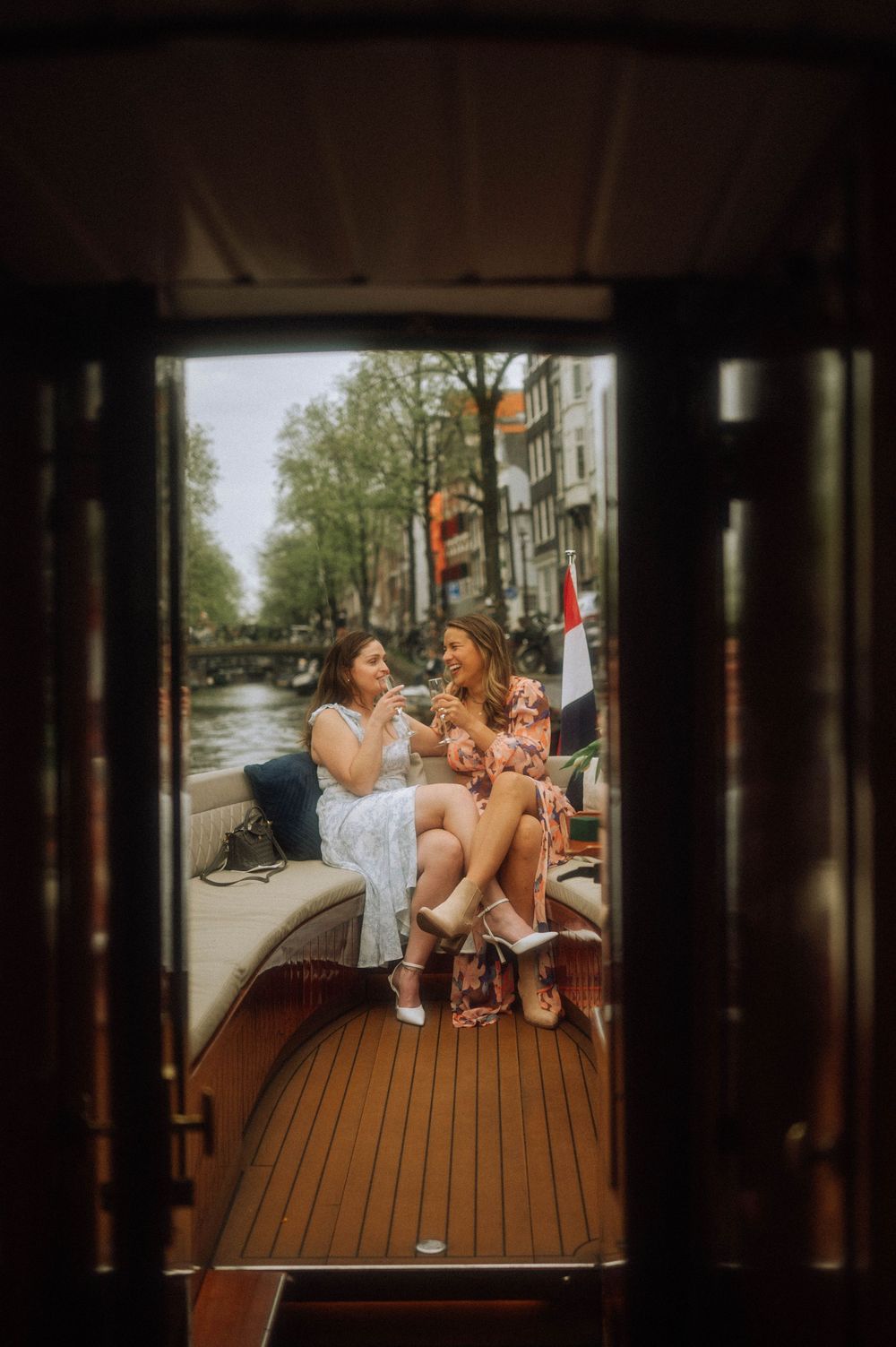 Two women sitting on a boat deck enjoying drinks on an Amsterdam canal, viewed through a wooden doorframe.