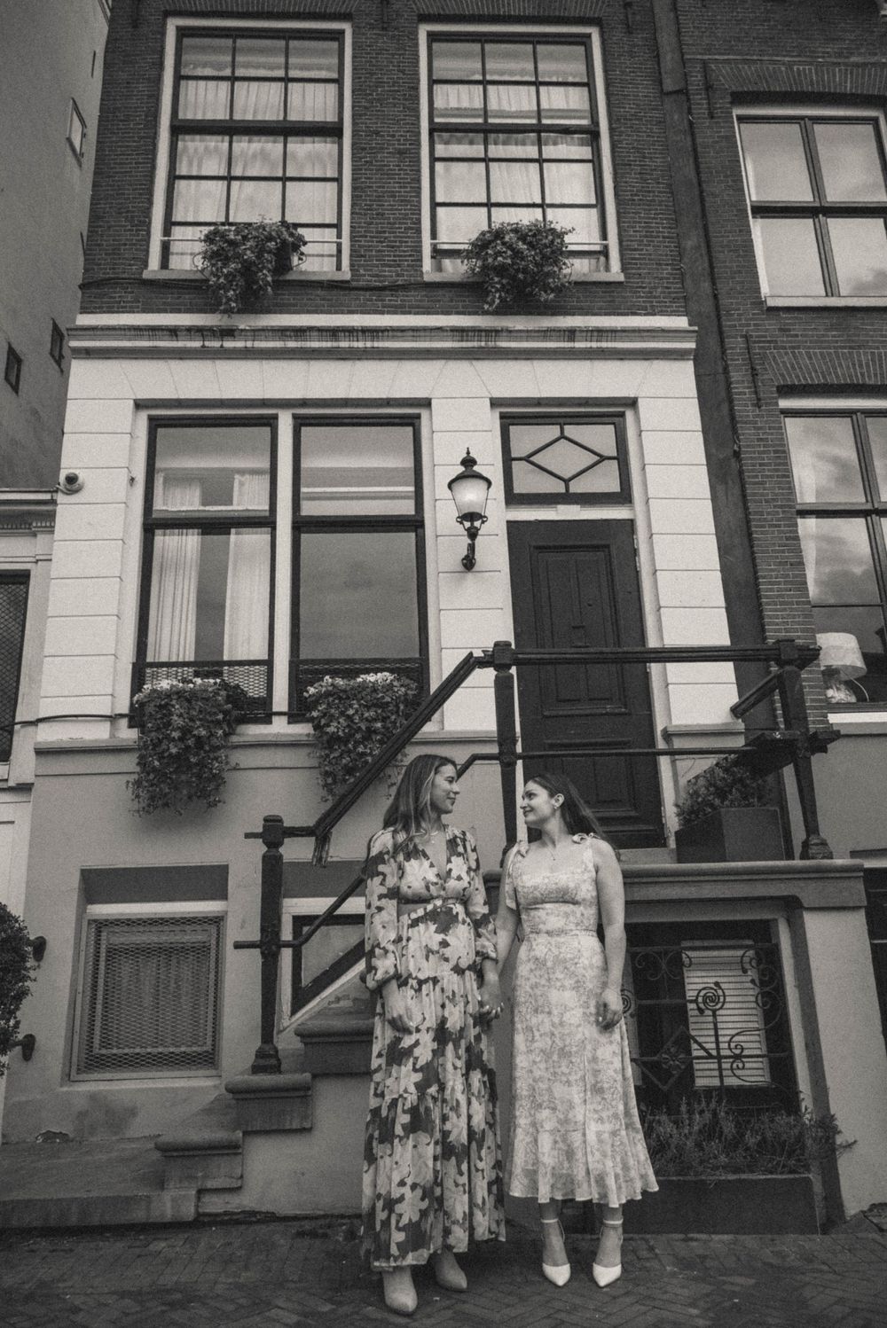 Two women in elegant dresses pose together in front of a classic Amsterdam canal house with flower boxes.