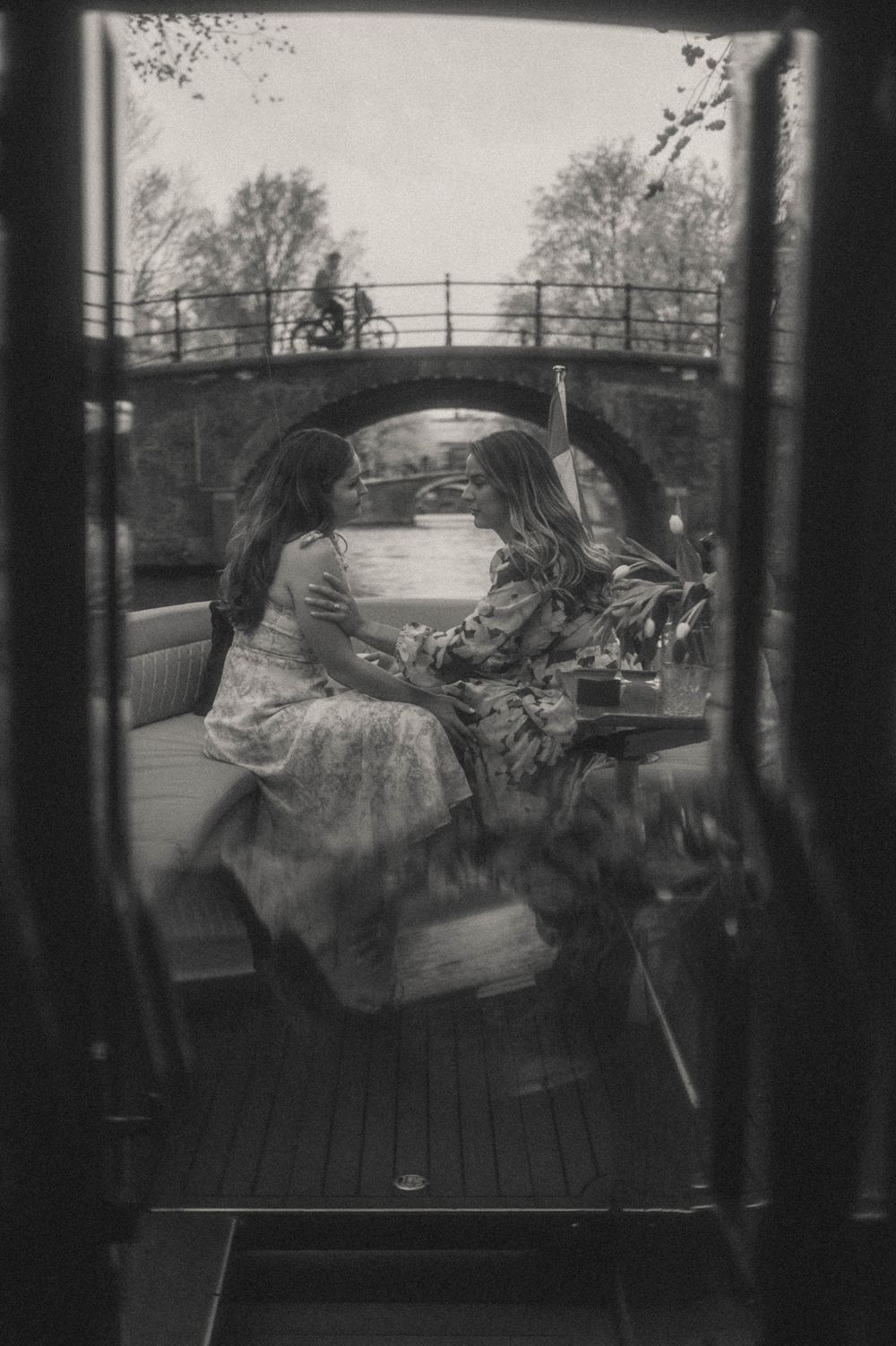 Two women sit together on a canal boat in Amsterdam, captured in black and white through a window.