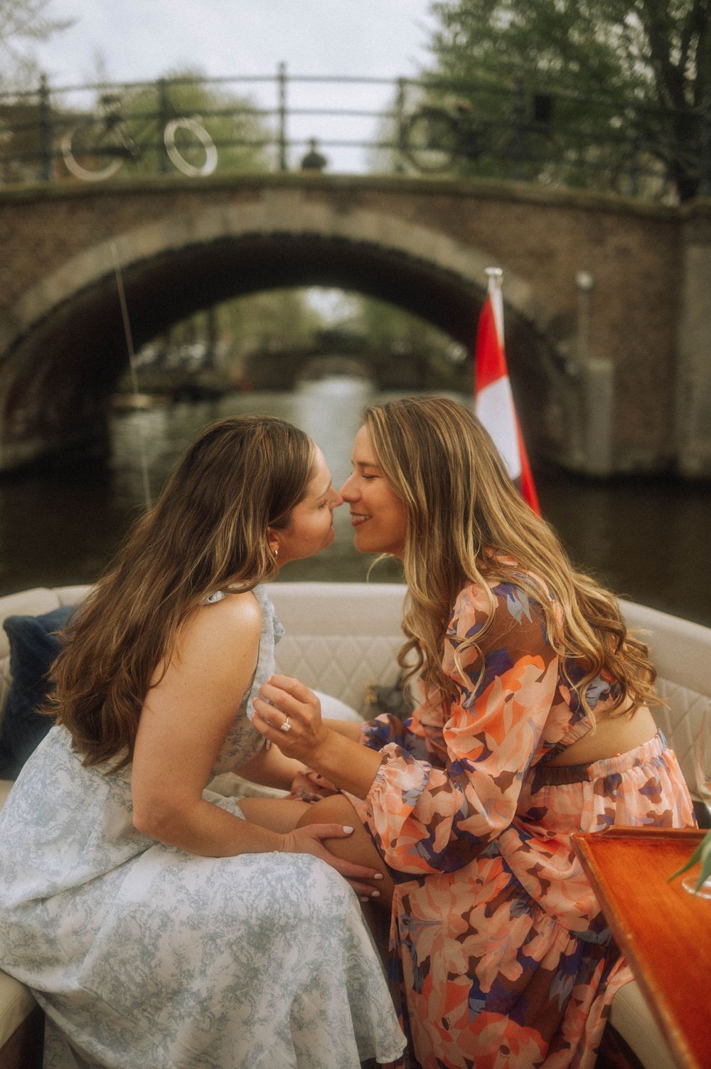 Two women share a romantic moment on a canal boat in Amsterdam, with a historic bridge and Dutch flag in the background.