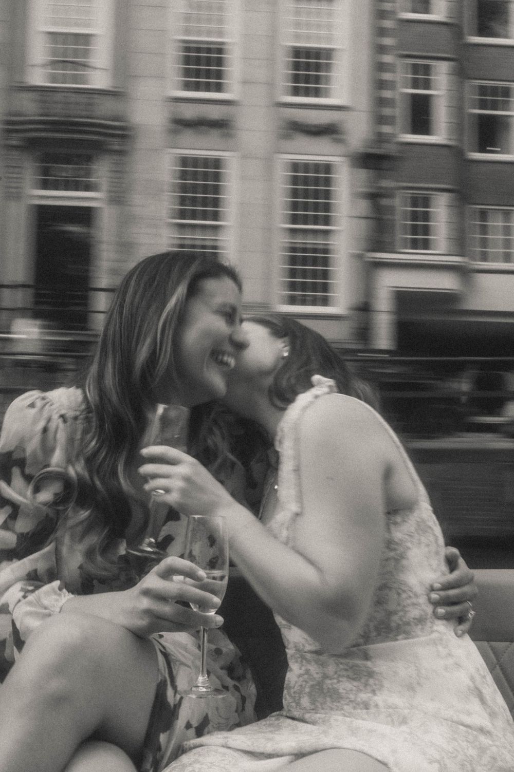 Two women laughing and sharing a kiss while holding champagne glasses on a boat in Amsterdam.