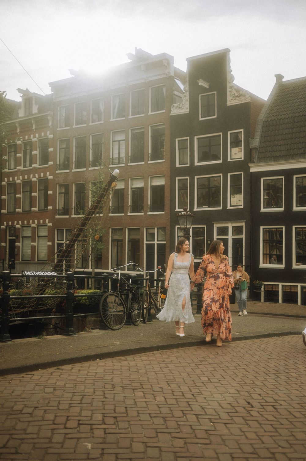 Two women in floral dresses walk hand-in-hand along Amsterdam canal cobblestone street with historic Dutch buildings backdrop.