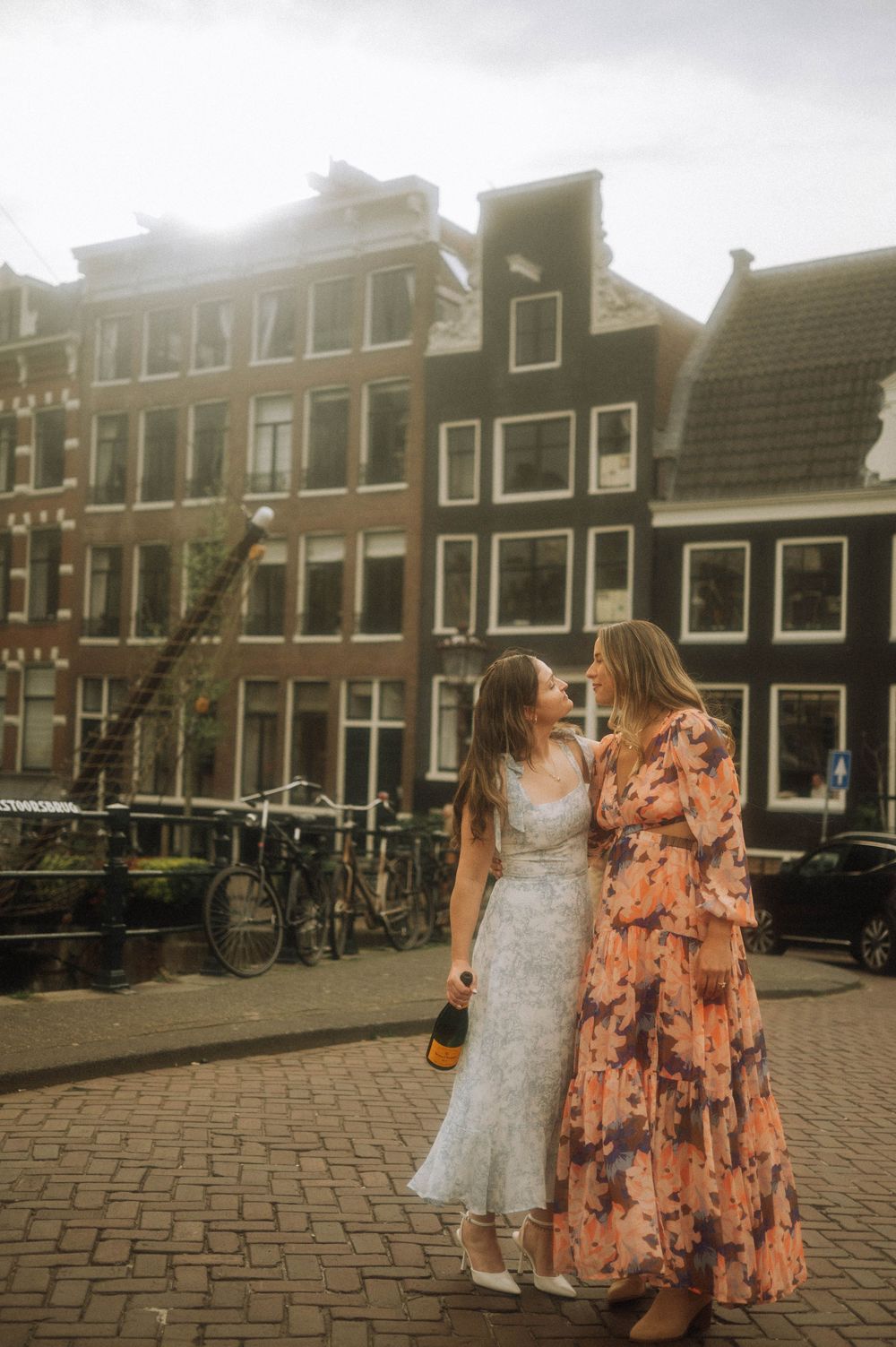 Two women in elegant dresses stand together on Amsterdam canal street, with historic Dutch buildings in the background.