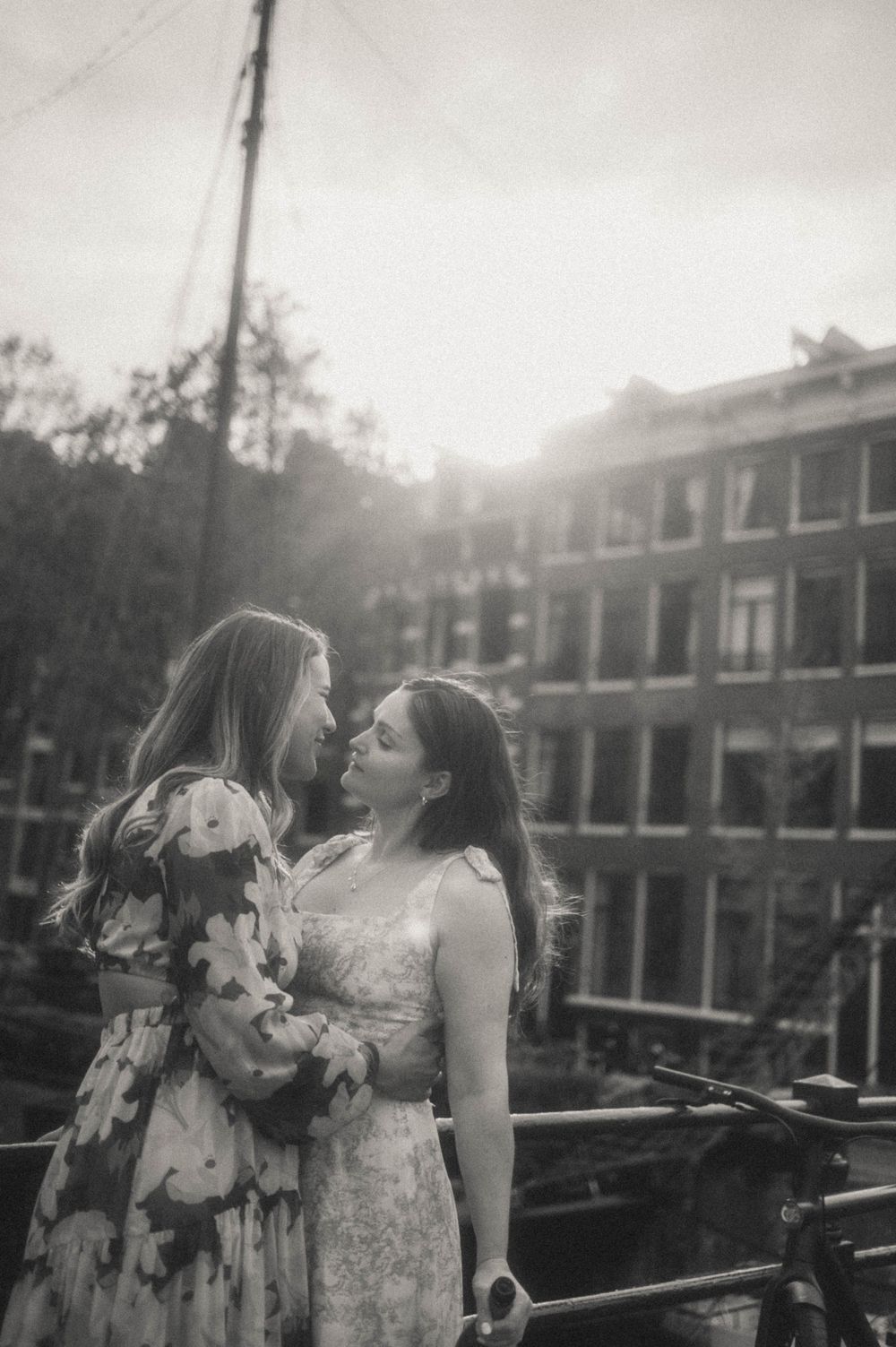 Two women in floral dresses share a romantic moment on a bridge in Amsterdam, backlit by golden sunlight.