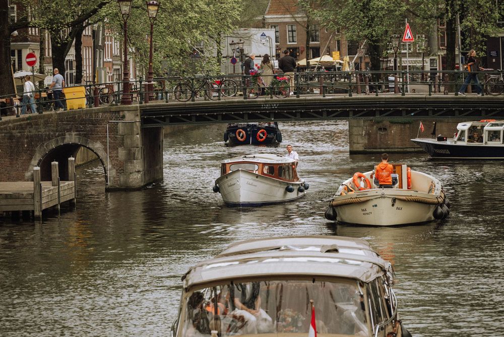 Boats navigating a busy Amsterdam canal under a stone bridge surrounded by lush trees.