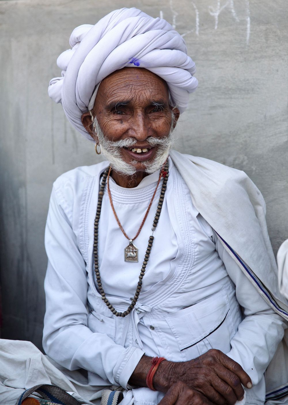Elderly Indian man in white turban and traditional attire wearing beaded necklaces, smiling warmly.
