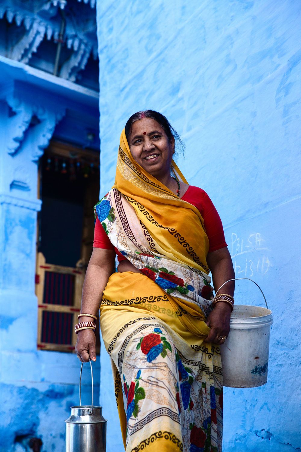 Smiling Indian woman in yellow floral sari carrying metal buckets against a vivid blue painted wall in Jodhpur.