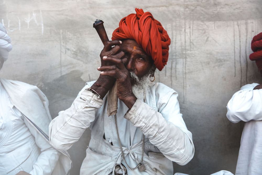 Elderly Indian man in white clothing and red turban smoking a traditional clay pipe at an outdoor gathering.