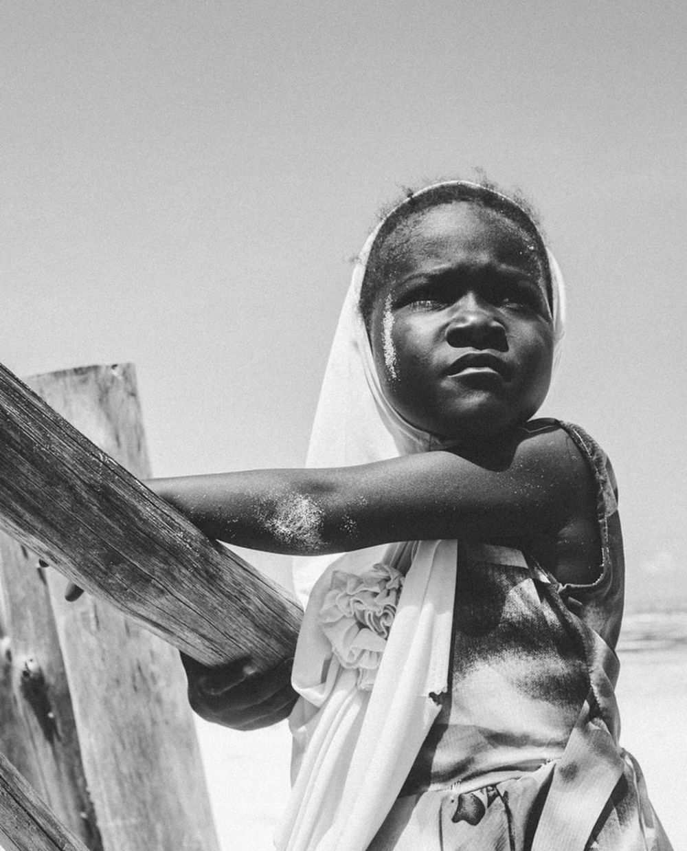 Black and white portrait of a young African child leaning on wooden posts under bright sky.