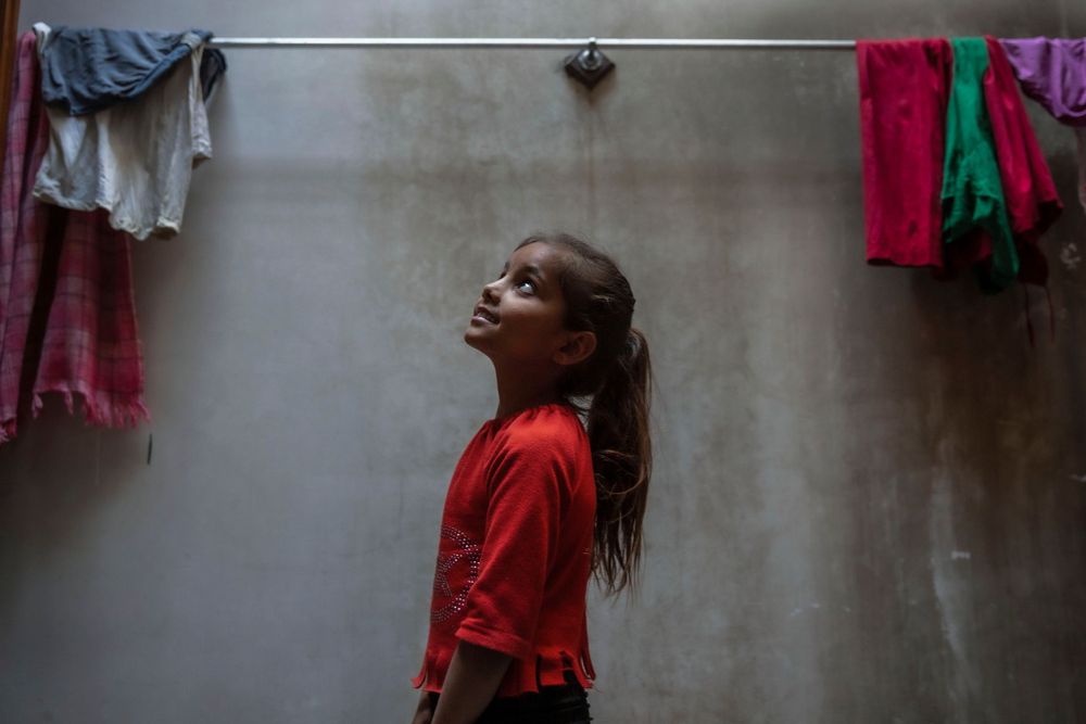 Young girl in red shirt standing under a shower head in a concrete bathroom, looking up.