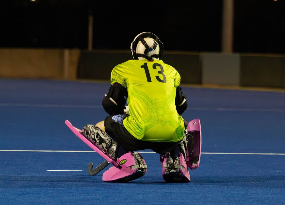 A field hockey goalkeeper in yellow jersey number 13 crouches with pink pads on blue turf.