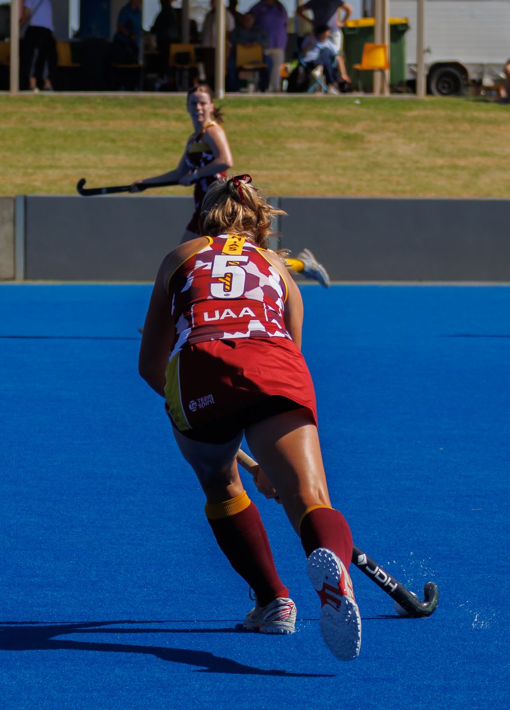 A female field hockey player in red UAA uniform sprints across a blue turf field.