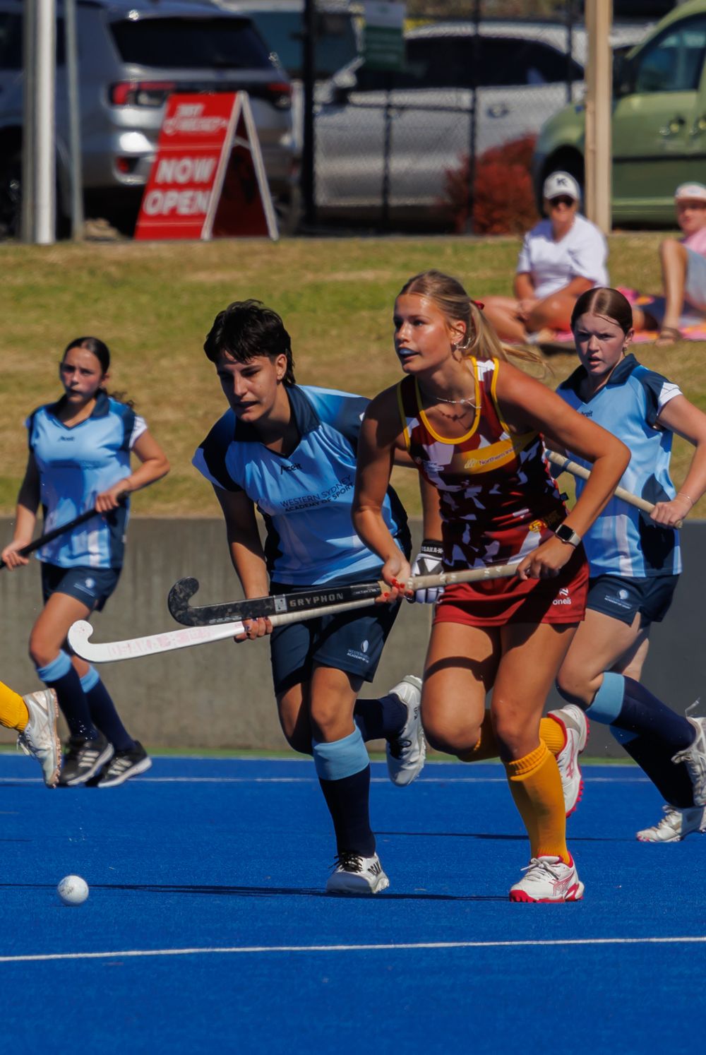 Female field hockey players compete on a blue turf court during an outdoor match.