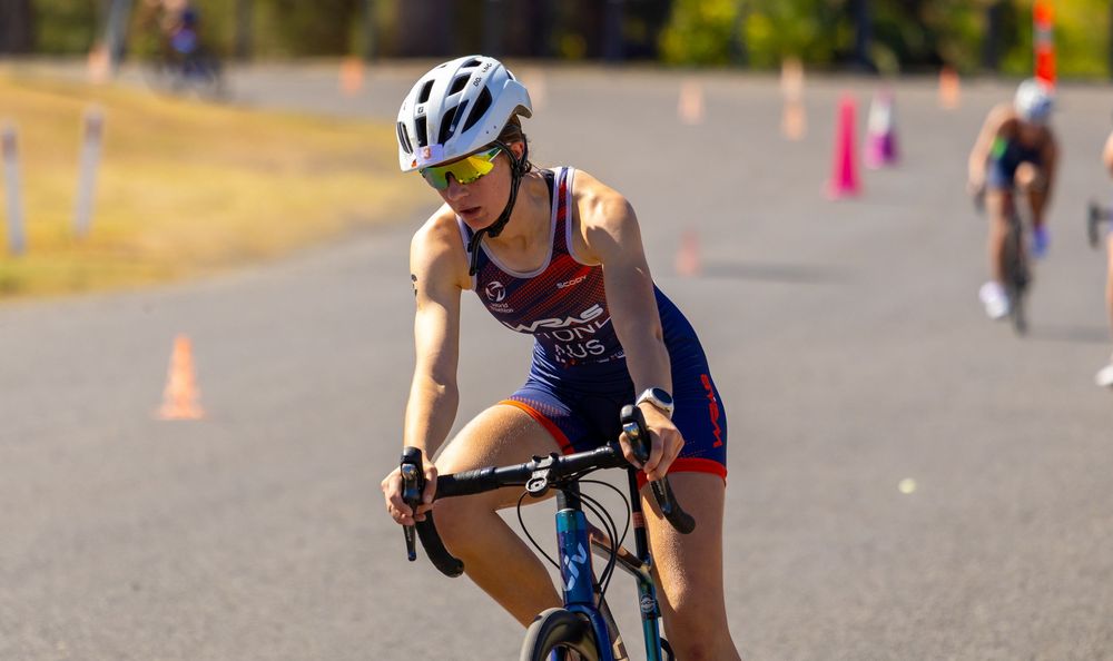Female triathlete in blue and red kit cycling on a road course with orange cones visible.