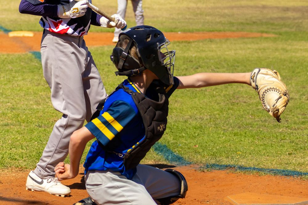Youth baseball catcher in blue uniform throwing ball during a game with batter in background.