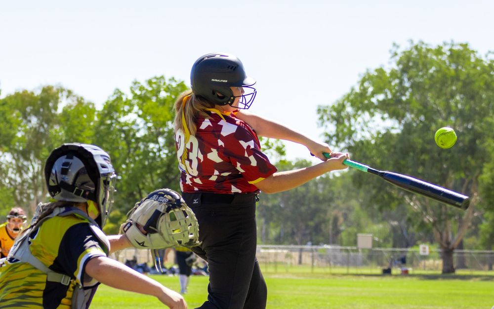 A female softball player swings a bat at a yellow ball during a competitive game outdoors.