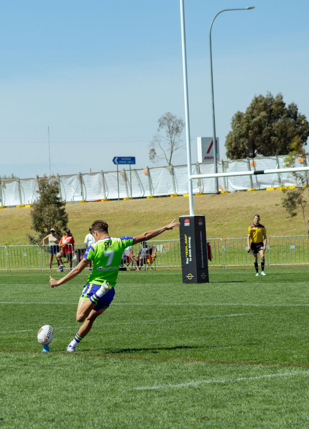 A rugby league player kicks a ball near a goal post on a sunny green field.