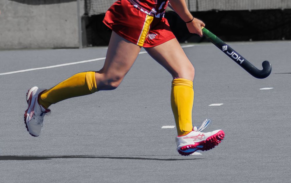 Female field hockey player in red uniform with yellow socks mid-stride on court holding a hockey stick.