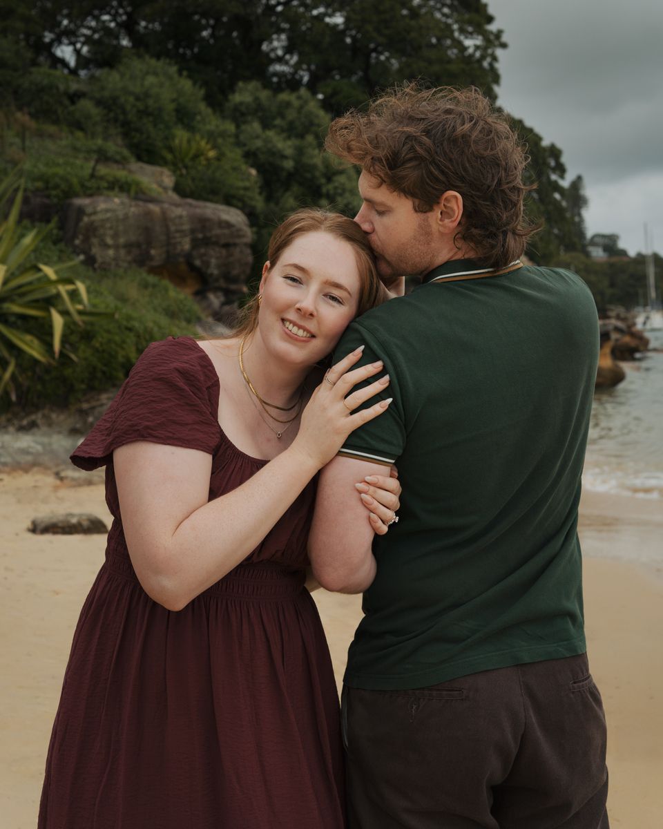 A couple hugs on a sandy beach, woman in burgundy dress smiling as man kisses her cheek near rocky shore during an engagement photoshoot