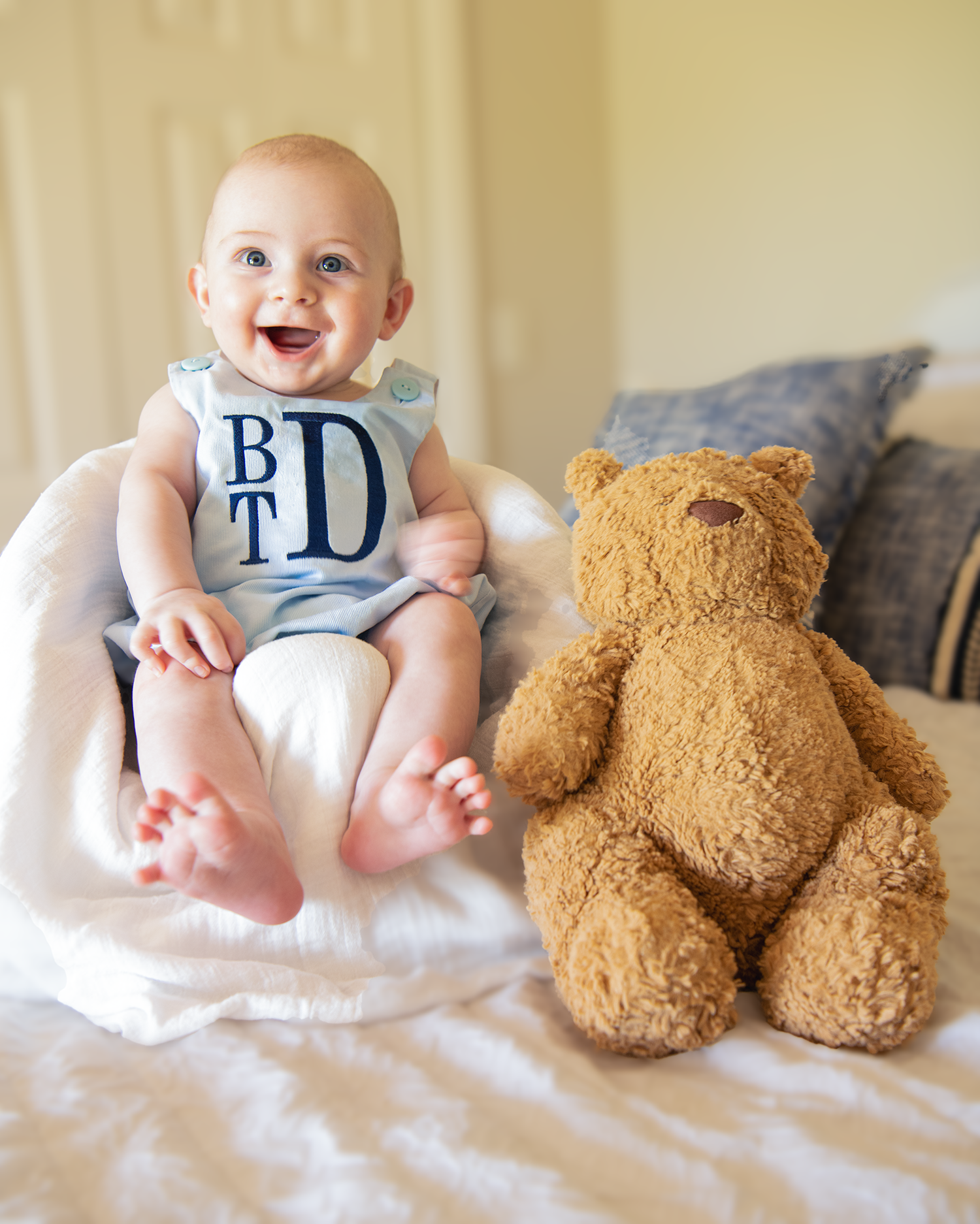 Happy baby in a monogrammed romper sits smiling beside a large teddy bear on a white bed.