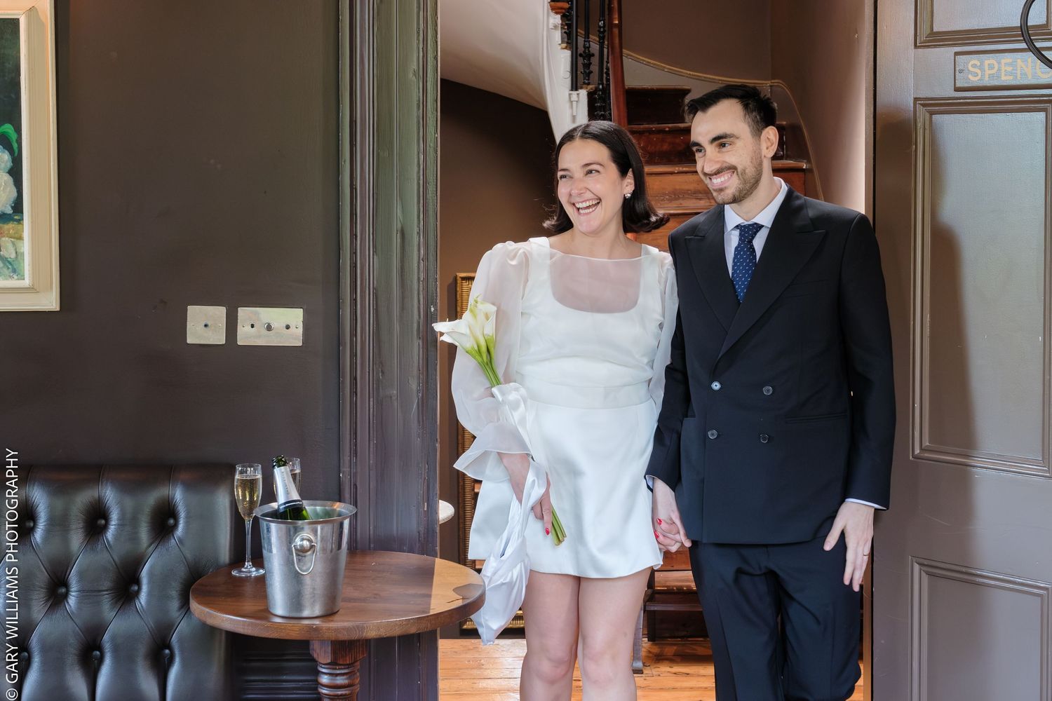 Smiling bride in white mini dress holding calla lily walks with groom in black suit at Canonbury Tavern