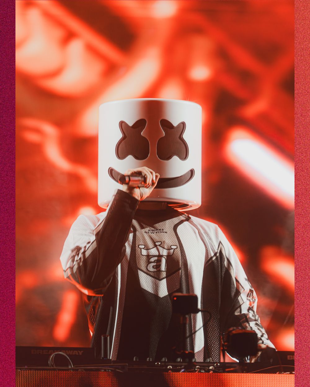 DJ Marshmello performing live on stage wearing iconic white helmet mask under dramatic red lighting.