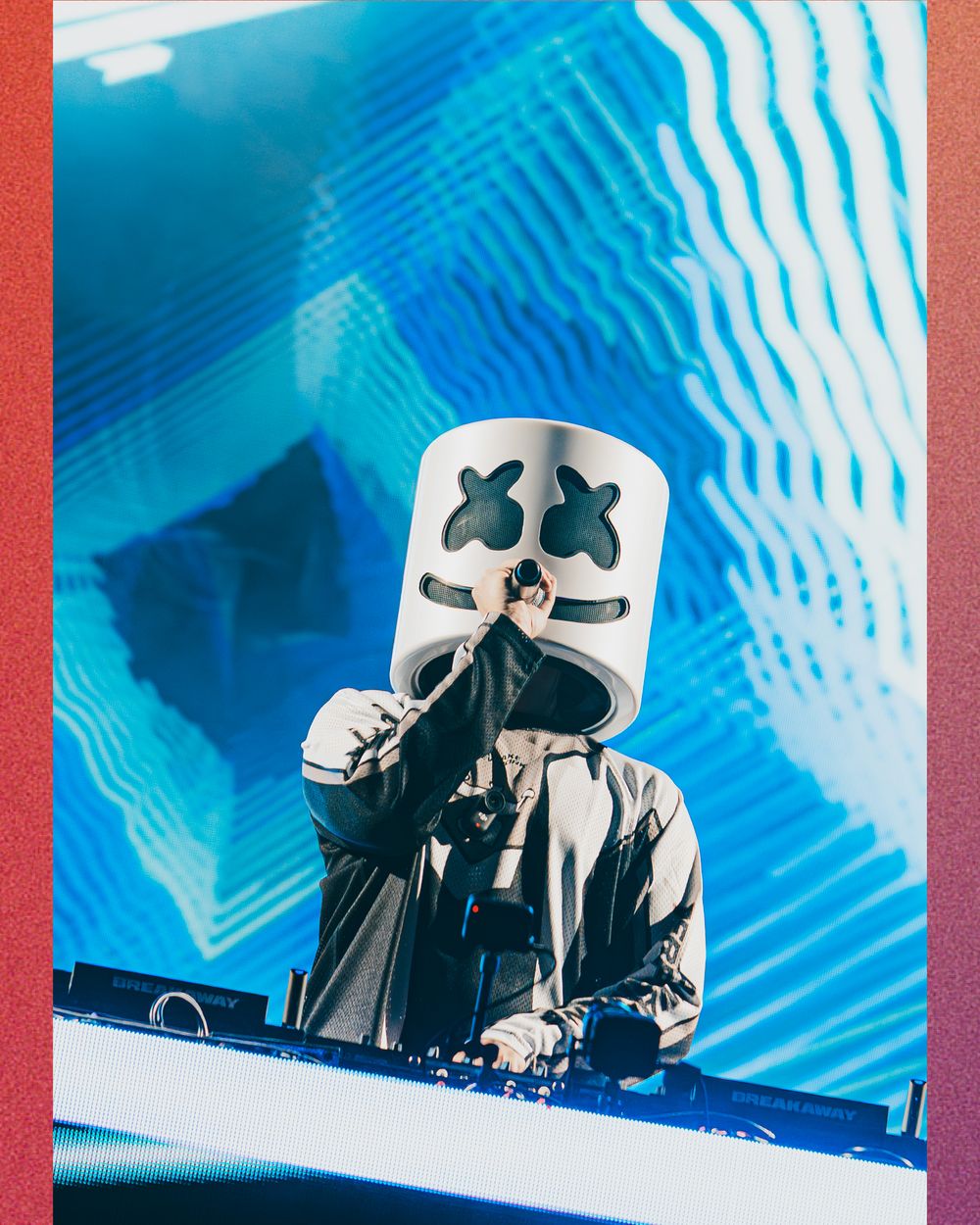 DJ Marshmello performing live on stage wearing iconic white helmet mask at a music festival with blue LED backdrop.
