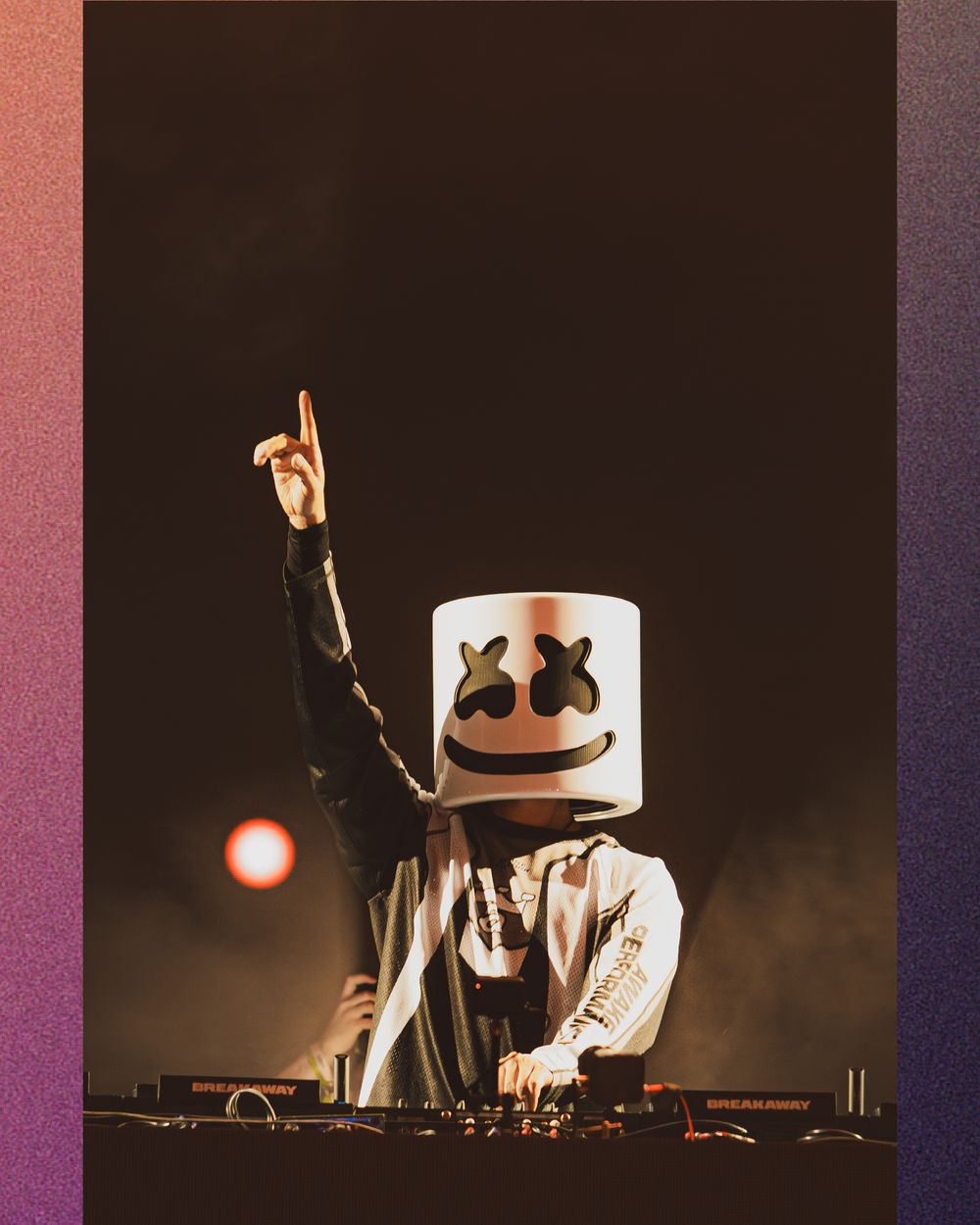 DJ Marshmello performing live on stage pointing finger up wearing signature white marshmallow helmet with crowd lights behind.