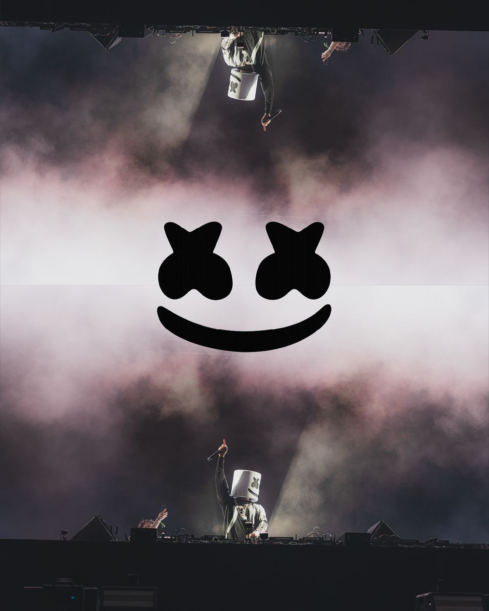 Marshmello DJ logo with smiley face over smoky concert stage performance with artist in white helmet.