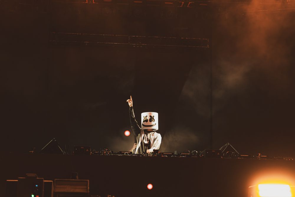 DJ Marshmello performing live on stage wearing iconic white helmet costume with dramatic lighting and fog effects.