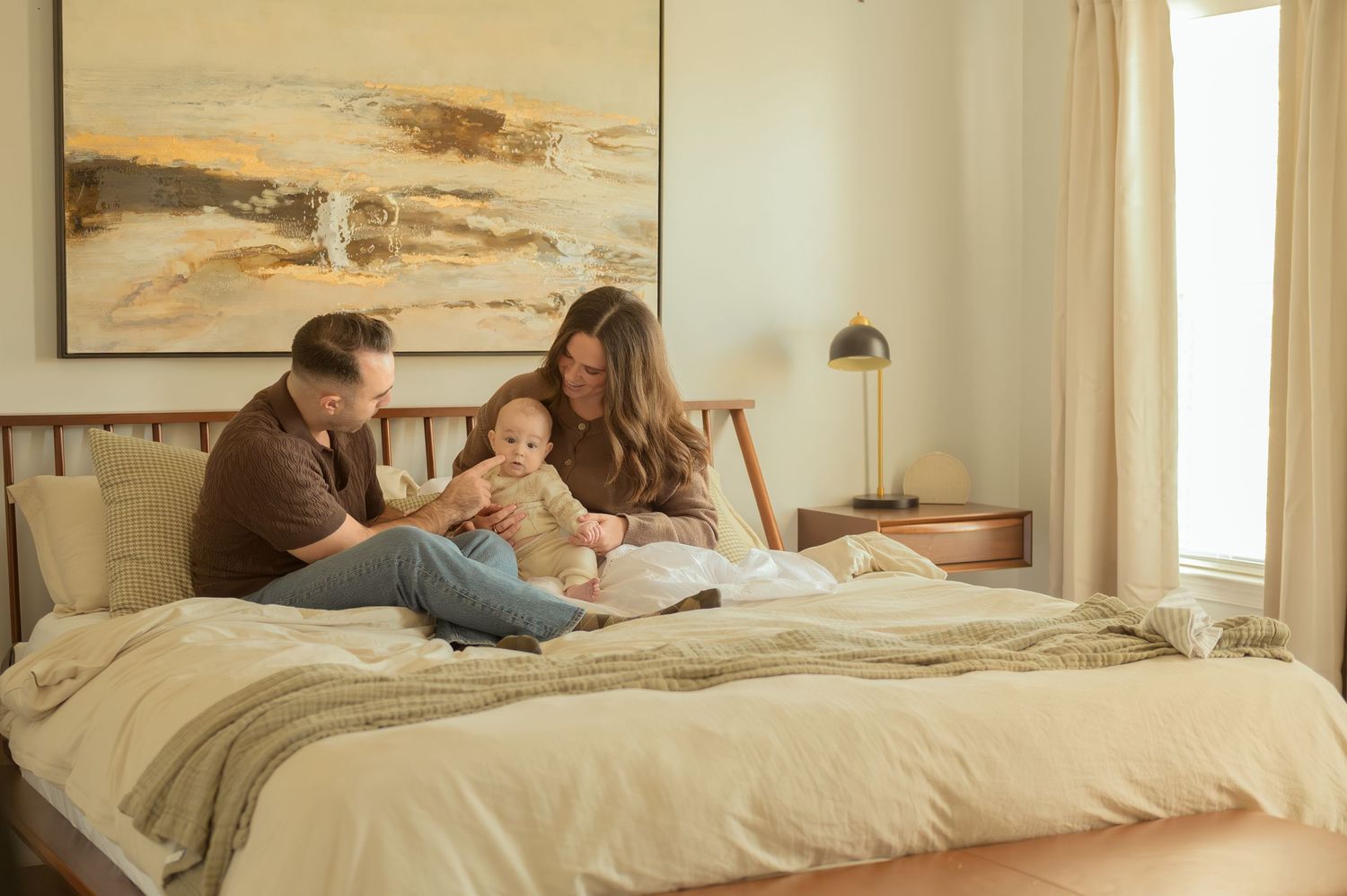 A young family sits on a bed, parents smiling at their newborn baby in a warmly decorated bedroom.