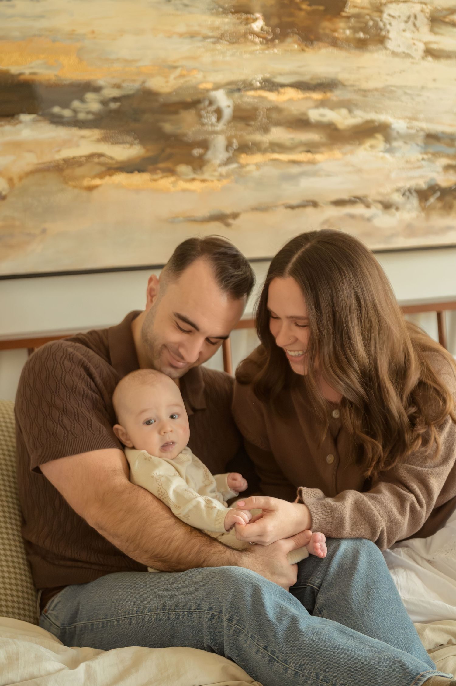 A couple sitting on a bed smiling at their newborn baby, with an abstract painting on the wall behind them.