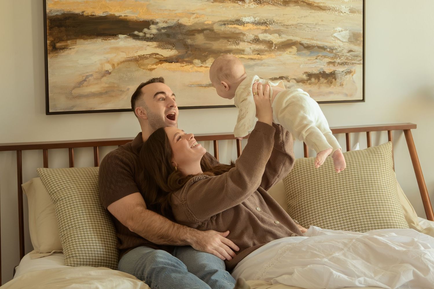 Happy parents sitting on bed laughing while mother holds smiling baby up in the air in cozy bedroom.