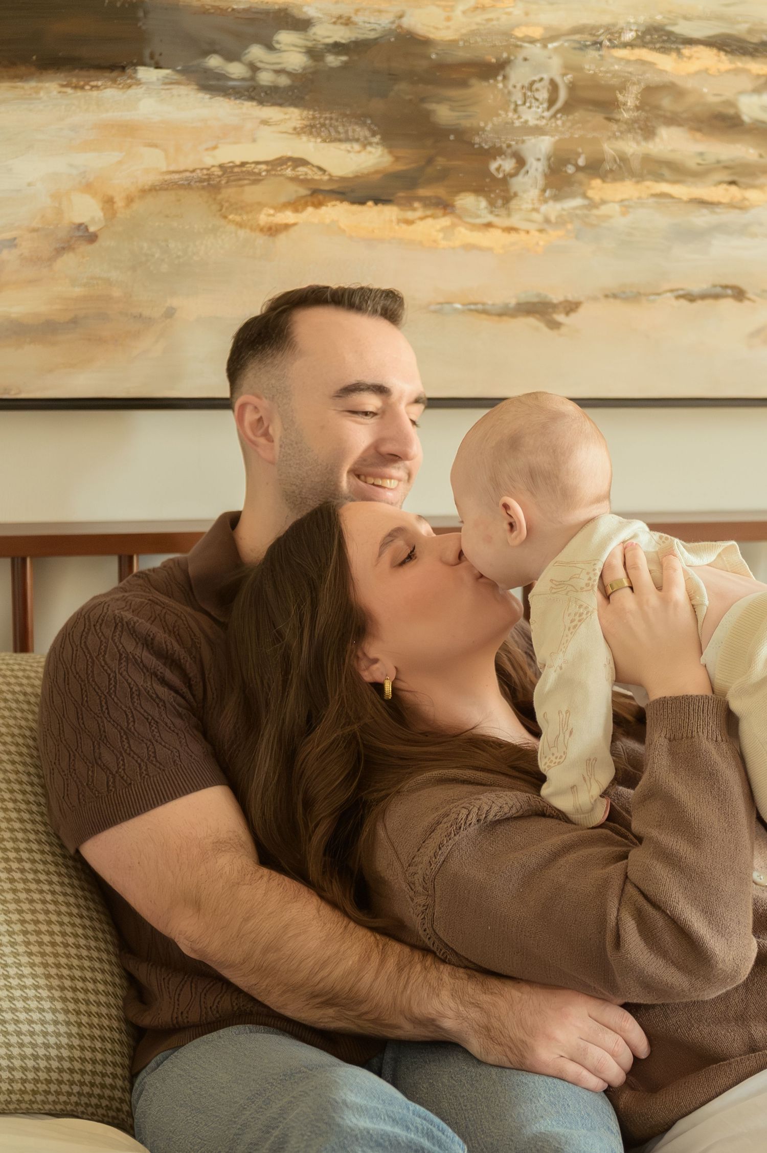 A smiling couple in brown sweaters sits on a couch, with the woman kissing their baby while the man looks on happily.