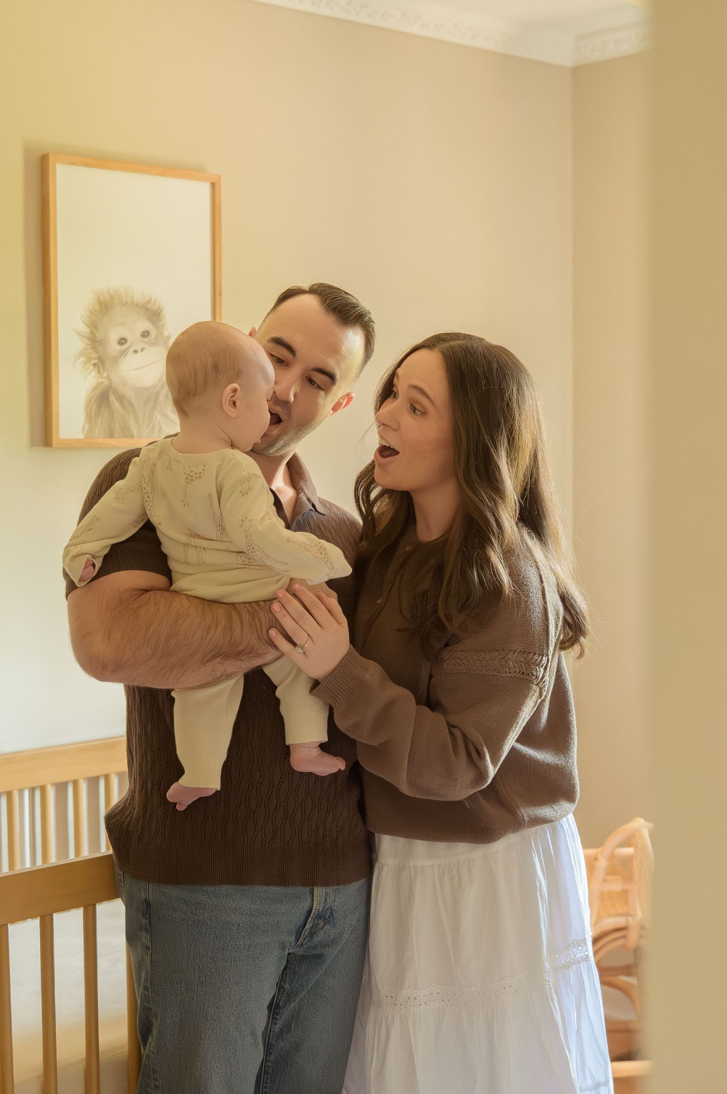 A couple in brown sweaters lovingly interact with their baby in a warmly decorated nursery room.