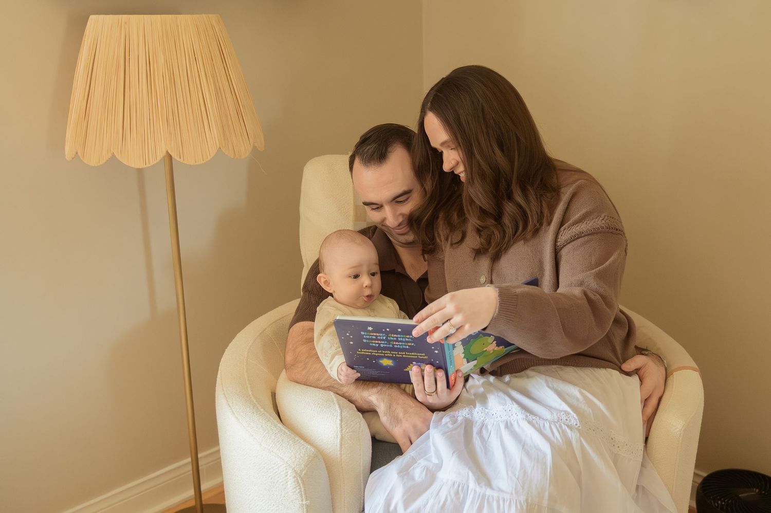Parents sitting in armchair reading a picture book to their newborn baby in a softly lit nursery.