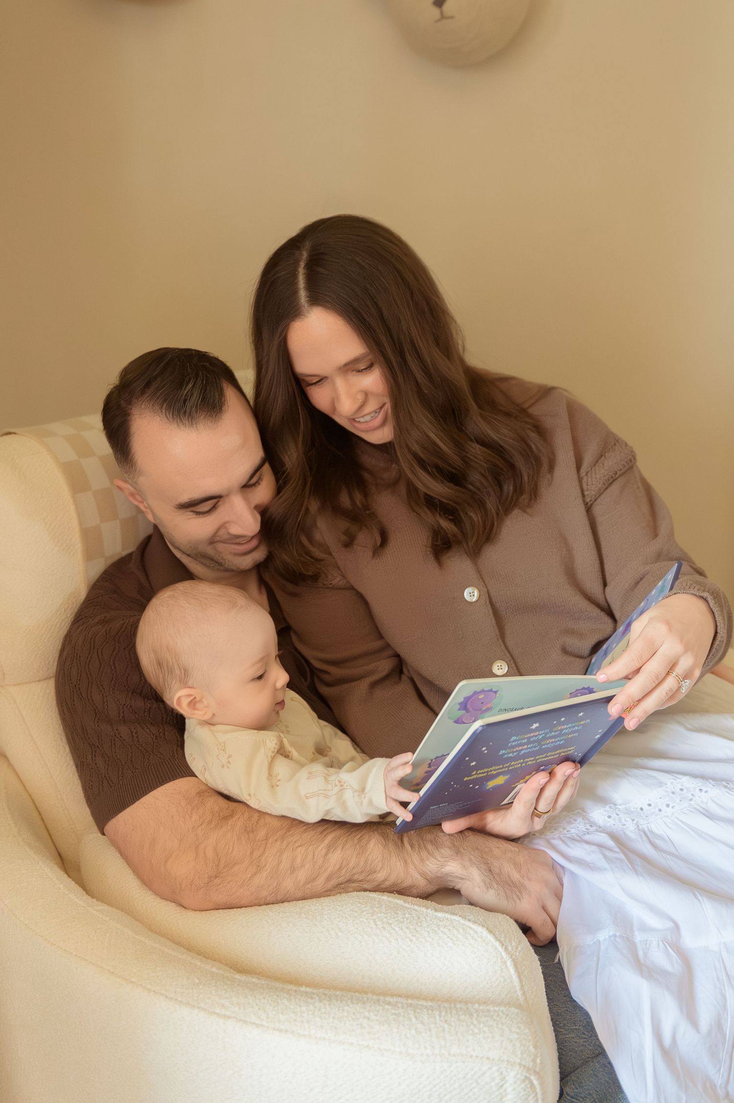 A happy family sits in a chair reading a picture book together with their baby in a cozy room.