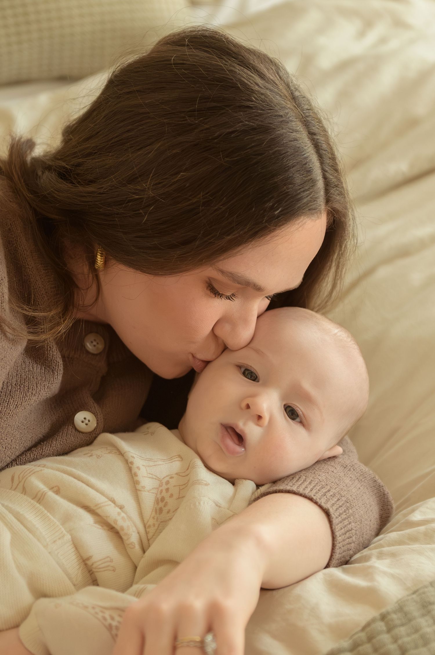 A loving mother gently kisses her newborn baby on the forehead while lying on a soft beige bed.
