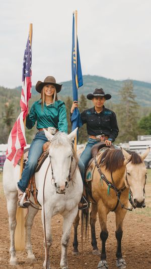 Two cowgirls on horseback holding American and state flags at a rodeo event.