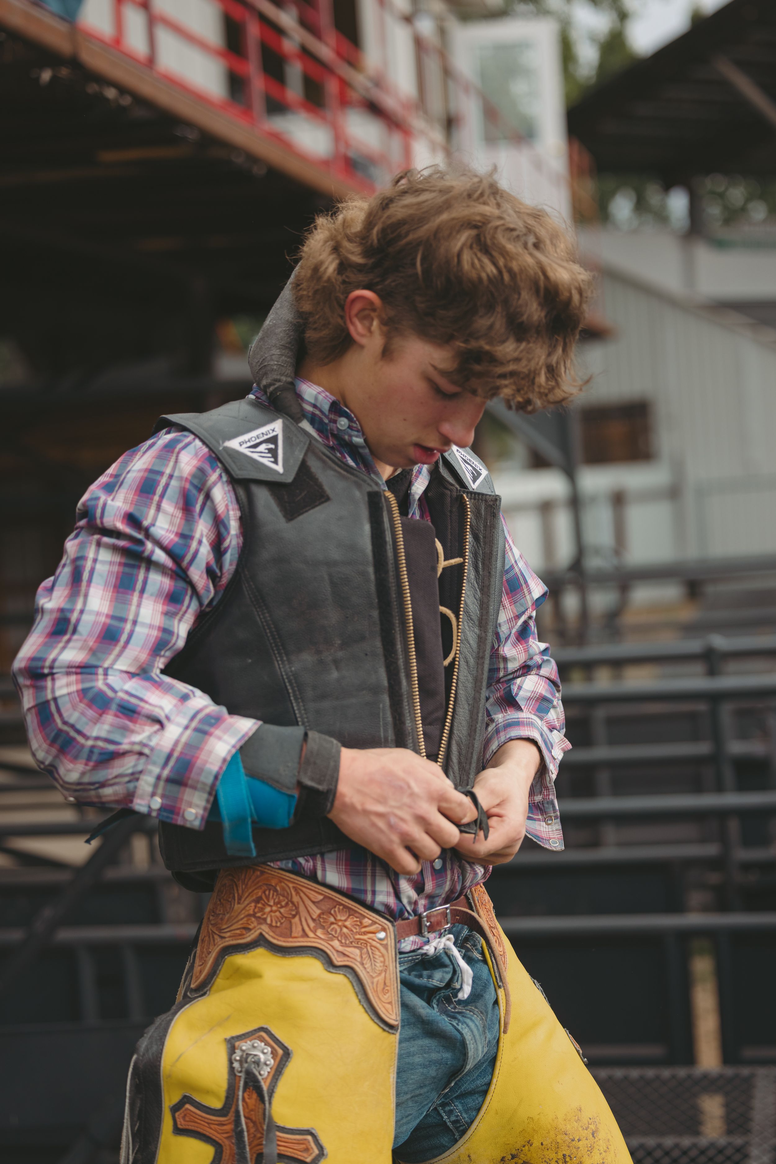 Young cowboy in plaid shirt and yellow chaps adjusting his gear at a rodeo event.
