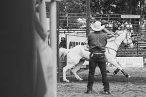Black and white photo of a cowboy watching a rider on a white horse in a rodeo arena.