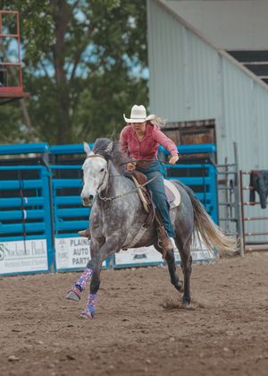 A cowgirl in a pink shirt races a dapple gray horse with colorful leg wraps at a rodeo event.