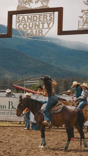 Cowgirl swinging a lasso on horseback under the Sanders County Fair sign at a rodeo.