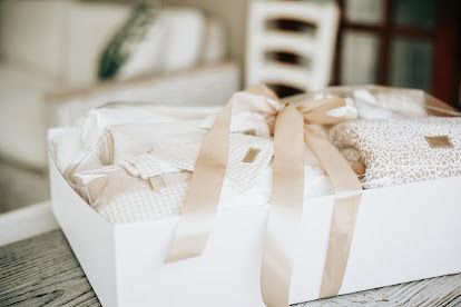 Elegant white gift box with satin ribbon bow, filled with neutral-toned baby items on a wooden table.