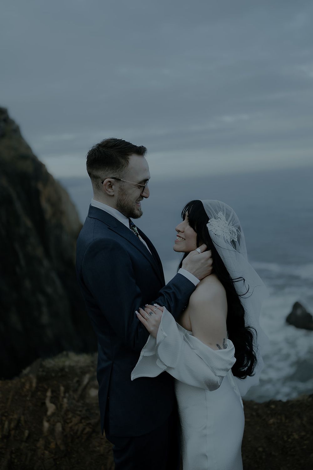 Couple embraces on a dramatic coastal cliff at dusk, bride in white gown with veil, groom in navy suit.