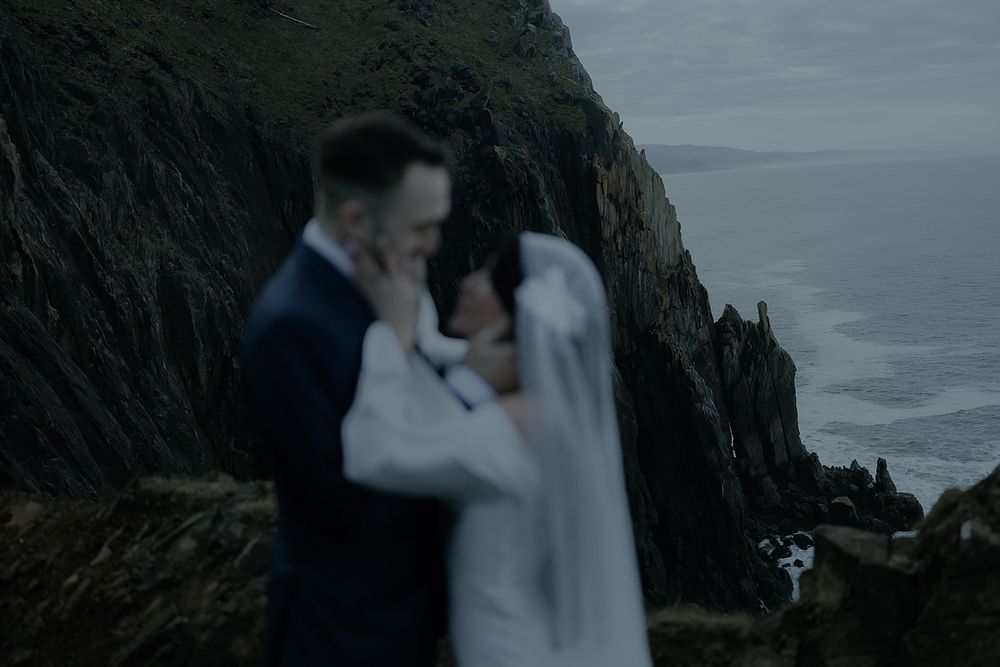 Couple sharing an intimate moment in wedding attire on dramatic coastal cliffs with ocean backdrop.