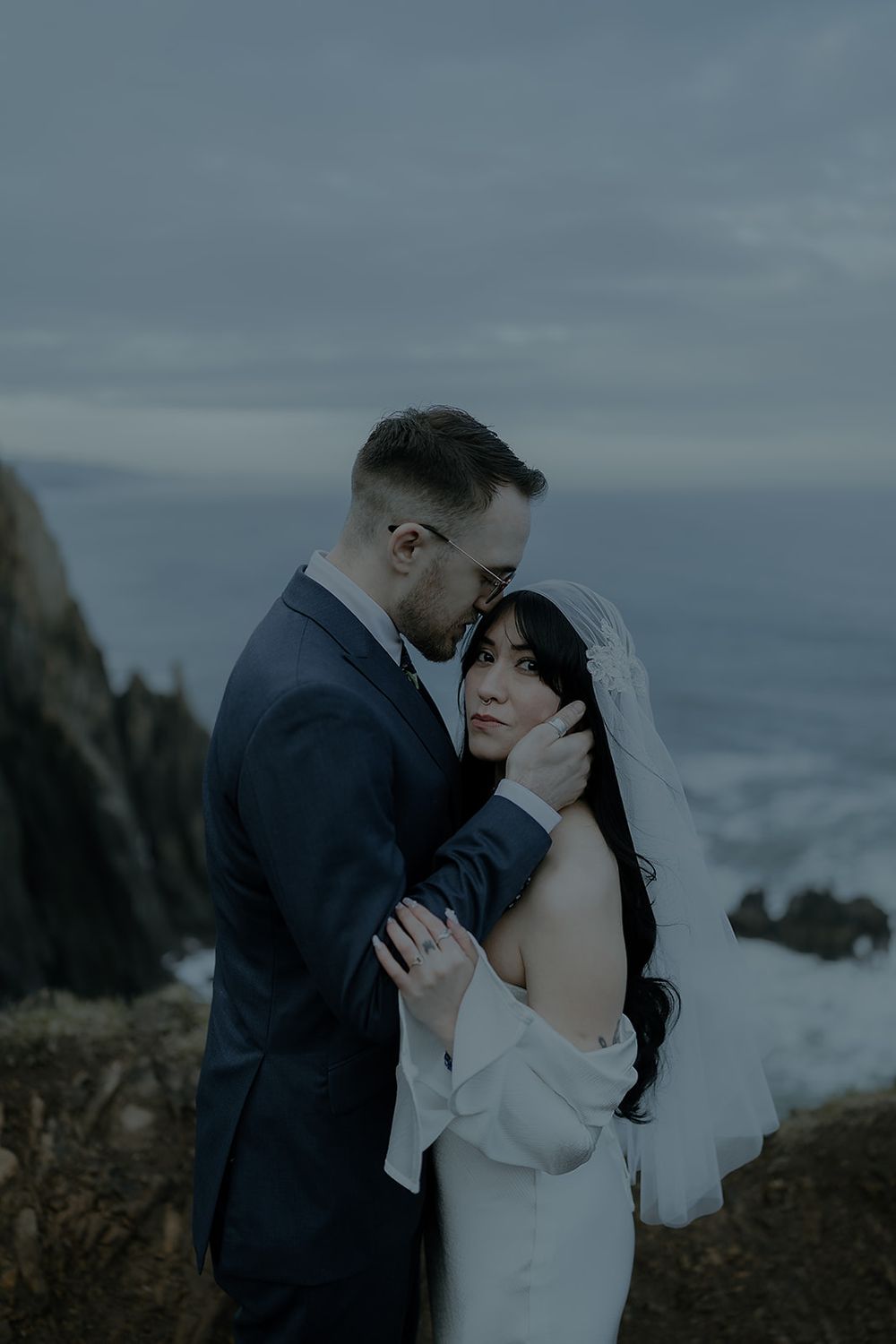 Couple embracing on coastal cliffs at dusk, bride in white gown with veil, groom in navy suit kissing her temple.
