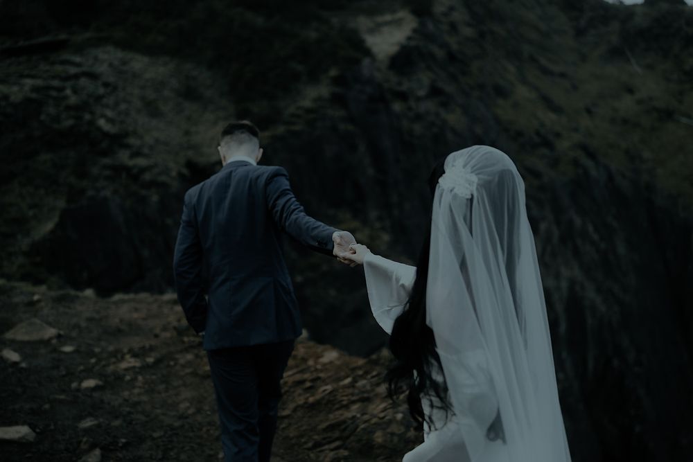 A couple walks hand-in-hand on rocky terrain, the bride wearing a flowing white veil, groom in dark suit.